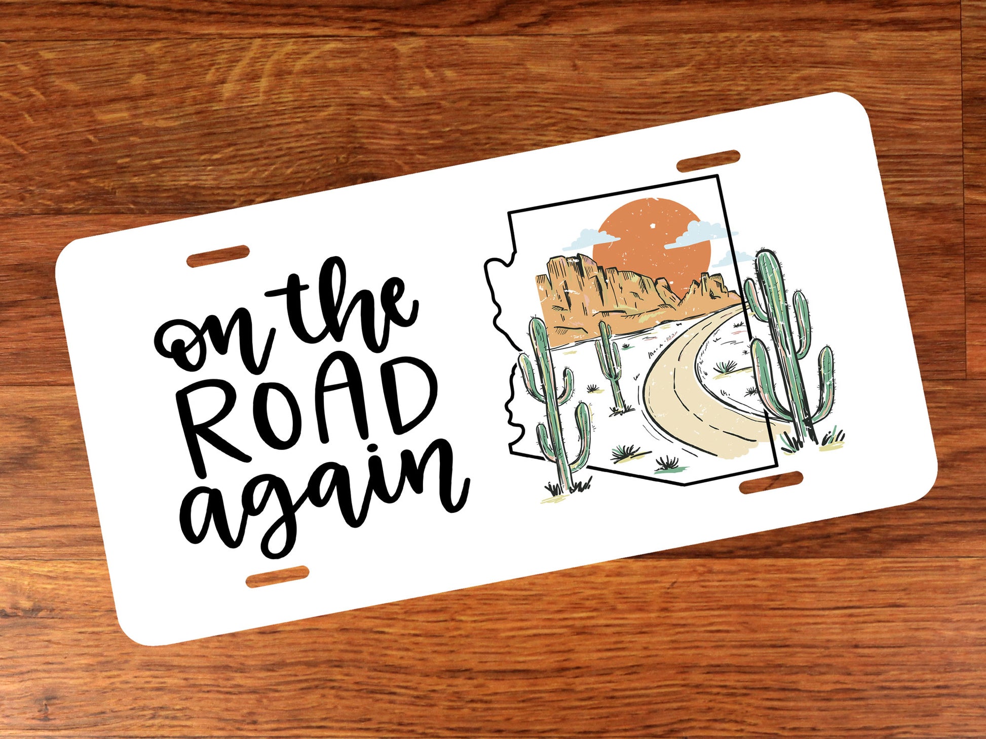 License plate with desert-themed design inside outline of Arizona state and 'on the road again' text on a wooden surface