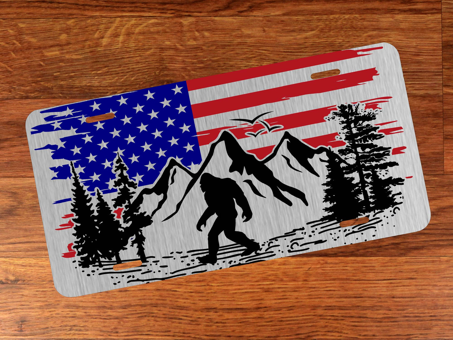 Silver license plate with American flag design featuring mountains, trees, and a Sasquatch on a wooden surface.