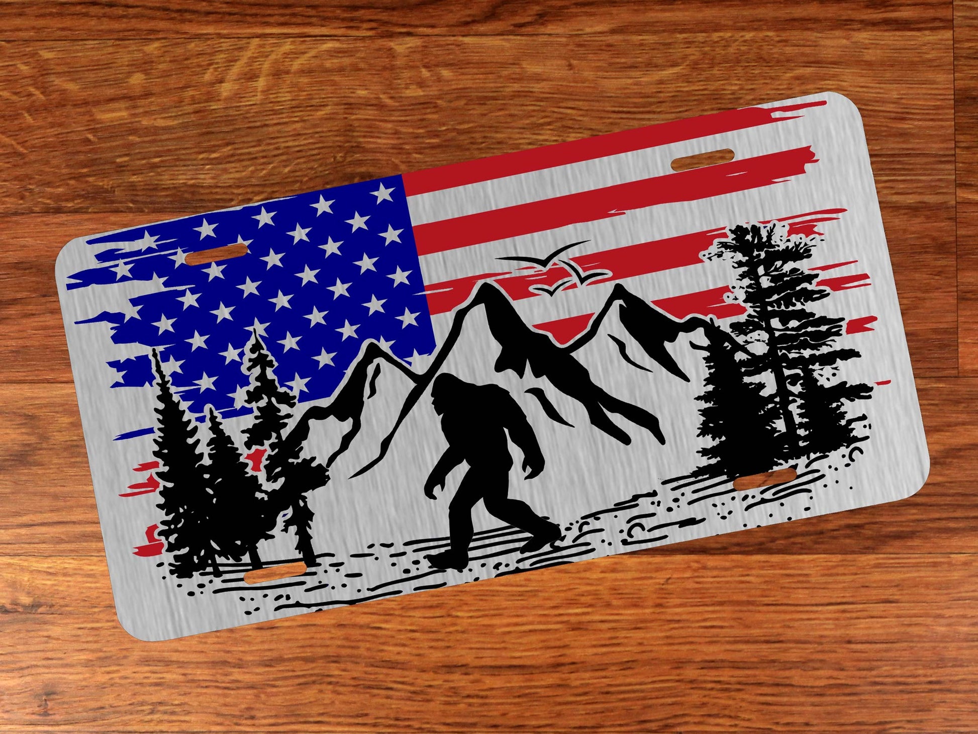 Silver license plate with American flag design featuring mountains, trees, and a Sasquatch on a wooden surface.