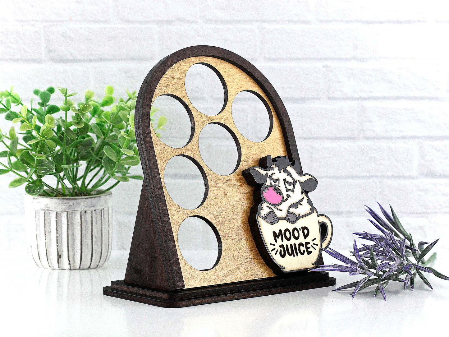 Wooden coffee pod display with a cow design and 'Mood Juice' text, placed on a white surface with plants.