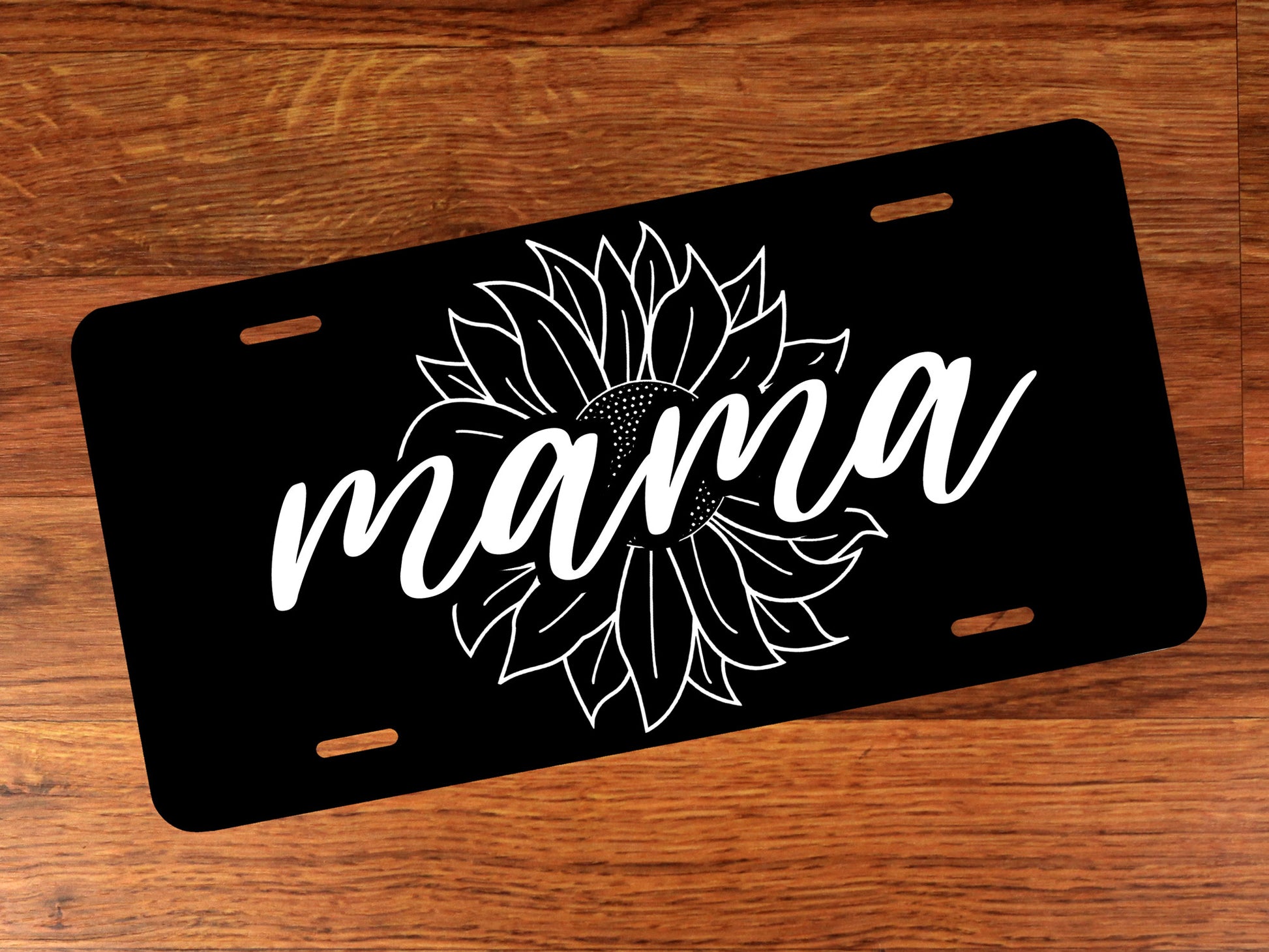 Black license plate with white 'mama' and sunflower design on a wooden surface